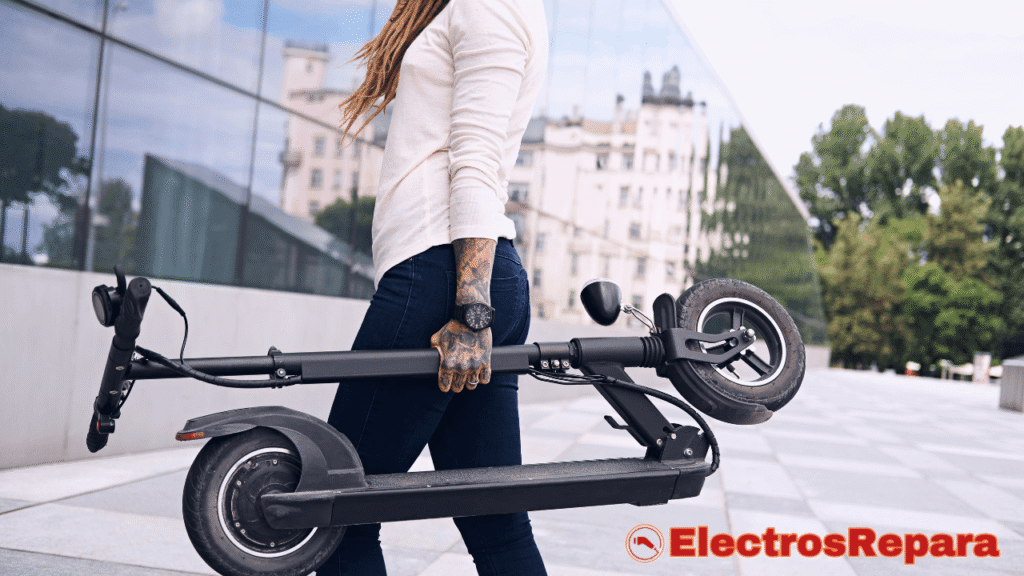 La imagen muestra a una persona (posiblemente una mujer con rastas y tatuajes en el brazo) caminando y llevando un patinete eléctrico plegado. El patinete es de color oscuro y parece estar en buen estado. El fondo sugiere un entorno urbano moderno con edificios de cristal y árboles. Aunque no es visible en la imagen, el Controlador de Patinete Eléctrico es un componente esencial de este tipo de vehículo, responsable de gestionar la potencia y las funciones del patinete que la persona transporta.