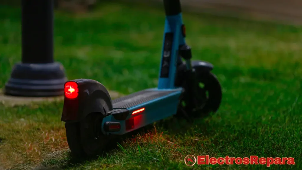 La imagen muestra un patinete eléctrico azul y negro con sus luces traseras encendidas, destacando las luces en patinetes eléctricos en un entorno de césped verde oscuro. La luz roja de la parte trasera del patinete y una tira de luz roja en el lateral están claramente visibles, lo que sugiere que el patinete está en funcionamiento o en modo de estacionamiento con visibilidad activada. En la parte inferior derecha de la imagen, se ve un logotipo con el texto "ElectrosRepara", lo que podría indicar la fuente de la imagen o el tema de la fotografía (reparación o mantenimiento de electrónica). La iluminación general de la escena es tenue, lo que resalta el brillo de las luces del patinete.