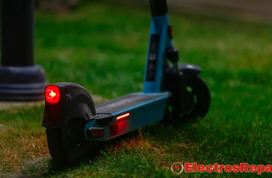 La imagen muestra un patinete eléctrico azul y negro con sus luces traseras encendidas, destacando las luces en patinetes eléctricos en un entorno de césped verde oscuro. La luz roja de la parte trasera del patinete y una tira de luz roja en el lateral están claramente visibles, lo que sugiere que el patinete está en funcionamiento o en modo de estacionamiento con visibilidad activada. En la parte inferior derecha de la imagen, se ve un logotipo con el texto "ElectrosRepara", lo que podría indicar la fuente de la imagen o el tema de la fotografía (reparación o mantenimiento de electrónica). La iluminación general de la escena es tenue, lo que resalta el brillo de las luces del patinete.