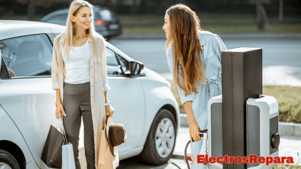 La imagen muestra a dos mujeres interactuando en un punto de carga de vehículos eléctricos. Una mujer de cabello largo y castaño está conectando o desconectando un cable del cargador, mientras la otra mujer, de cabello rubio, la mira y sonríe, sosteniendo bolsas de compra. Un coche eléctrico blanco está parcialmente visible detrás de ellas.