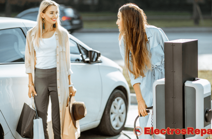 La imagen muestra a dos mujeres interactuando en un punto de carga de vehículos eléctricos. Una mujer de cabello largo y castaño está conectando o desconectando un cable del cargador, mientras la otra mujer, de cabello rubio, la mira y sonríe, sosteniendo bolsas de compra. Un coche eléctrico blanco está parcialmente visible detrás de ellas.