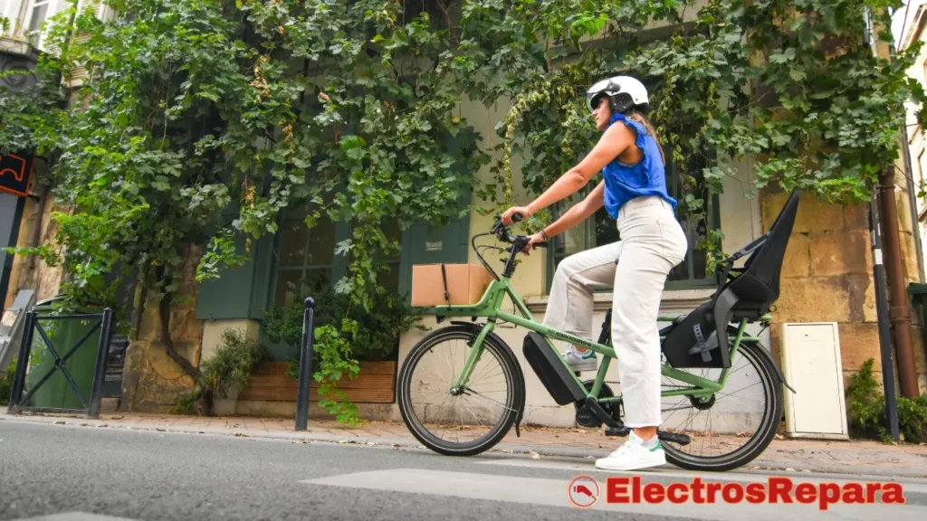 una mujer con casco conduciendo una bicicleta eléctrica de color verde por una calle de adoquines. La bicicleta tiene una caja en la parte delantera y un asiento para niños vacío en la parte trasera. El entorno parece urbano, con edificios cubiertos de vegetación a los lados de la calle. La mujer está pedaleando, lo que podría estar relacionado con el Pedaleo Intermitente en tu E-Bike.