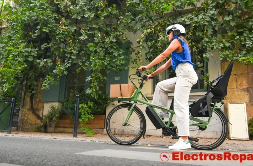 una mujer con casco conduciendo una bicicleta eléctrica de color verde por una calle de adoquines. La bicicleta tiene una caja en la parte delantera y un asiento para niños vacío en la parte trasera. El entorno parece urbano, con edificios cubiertos de vegetación a los lados de la calle. La mujer está pedaleando, lo que podría estar relacionado con el Pedaleo Intermitente en tu E-Bike.