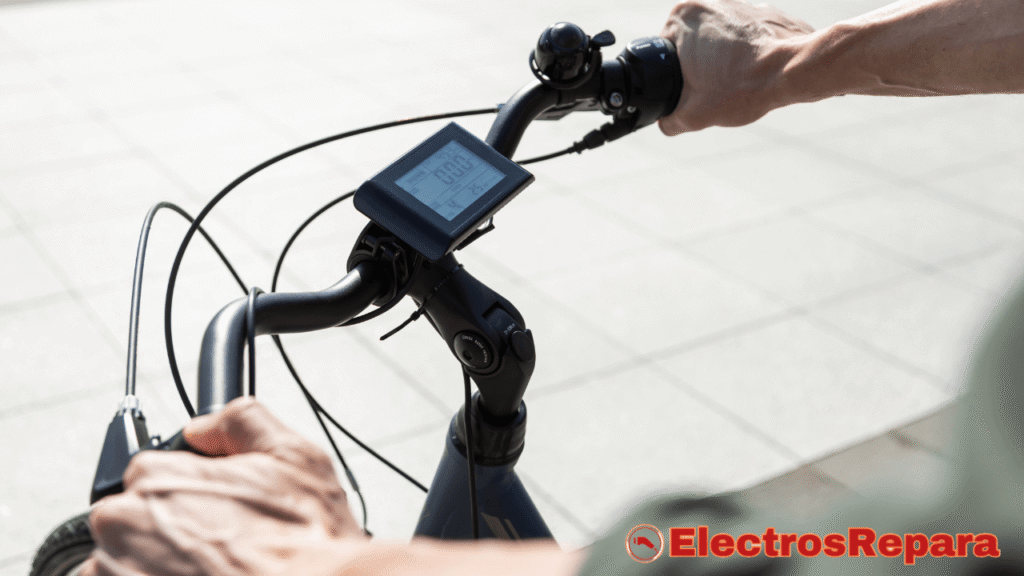 La imagen muestra un primer plano de las manos de una persona sosteniendo el manillar de una bicicleta eléctrica. En el centro del manillar, se ve una pantalla digital que muestra información como la velocidad (0.00) y el nivel de asistencia. También hay una pequeña campana en el lado izquierdo del manillar. El fondo es un camino o pavimento exterior, con la luz del sol cayendo sobre él.