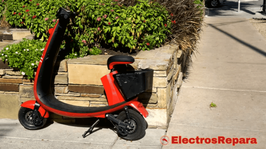 La imagen muestra un patinete eléctrico de color rojo brillante y negro, estacionado en una acera junto a un muro de piedra con vegetación frondosa en la parte superior. El patinete tiene un diseño robusto, con una cesta de almacenamiento en la parte trasera. Aunque las Baterías para Patinete Eléctrico no son visibles externamente, son el componente esencial que permite el funcionamiento de este vehículo, lo que lo convierte en una ilustración adecuada para un contexto relacionado con patinetes eléctricos y su energía.