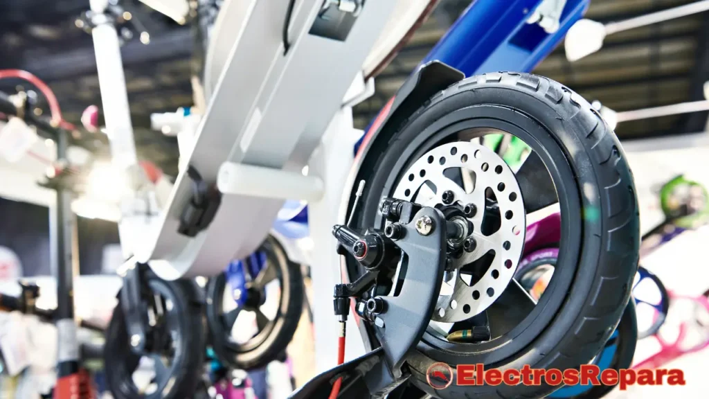 La imagen muestra la rueda trasera de un patinete eléctrico en primer plano, con un enfoque claro en el disco de freno y el neumático. La rueda parece estar en buenas condiciones, sin signos visibles de un pinchazo de rueda de patinete. En el fondo, se aprecian otros patinetes, lo que sugiere un entorno de tienda o exposición. La iluminación es brillante y resalta los detalles del patinete.