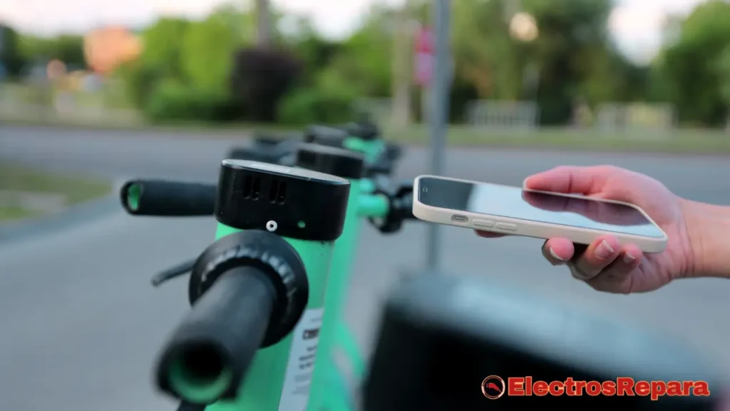 La imagen muestra la mano de una persona sosteniendo un smartphone, apuntándolo hacia el manillar de un patinete eléctrico de color verde brillante. El smartphone tiene una funda de color claro y la pantalla no es claramente visible para confirmar la aplicación. Sin embargo, en el contexto de la conectividad de patinetes, la persona podría estar intentando interactuar con la App Mi Home u otra aplicación de gestión de patinetes para activar o desbloquear el vehículo. La zona de la pantalla del patinete, que es negra, es visible en la parte superior del manillar. Al fondo, otros patinetes verdes aparecen difuminados, y se percibe follaje que sugiere un entorno exterior, posiblemente urbano o un parque.