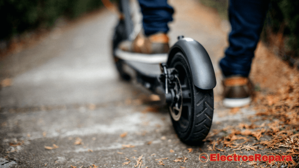 La imagen muestra una toma desde atrás de un patinete eléctrico en movimiento.
Se enfoca en la rueda trasera, el guardabarros y la plataforma del patinete. Se pueden ver las piernas de una persona que lo está conduciendo, con pantalones vaqueros y zapatos de color marrón claro.
