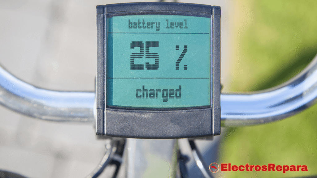 La imagen muestra una vista de primer plano de un monitor digital en el manillar de una bicicleta eléctrica. La pantalla LCD retroiluminada en color verde azulado es el foco principal, y muestra el estado de la batería. Las palabras "battery level" están en la parte superior, y el número grande "25%" está en el centro, indicando que la batería de la bicicleta eléctrica está a un cuarto de su carga total. Debajo del porcentaje, se puede leer la palabra "charged". La unidad de visualización está montada en un manillar cromado o de metal pulido, con la imagen de la bicicleta desenfocada en el fondo.