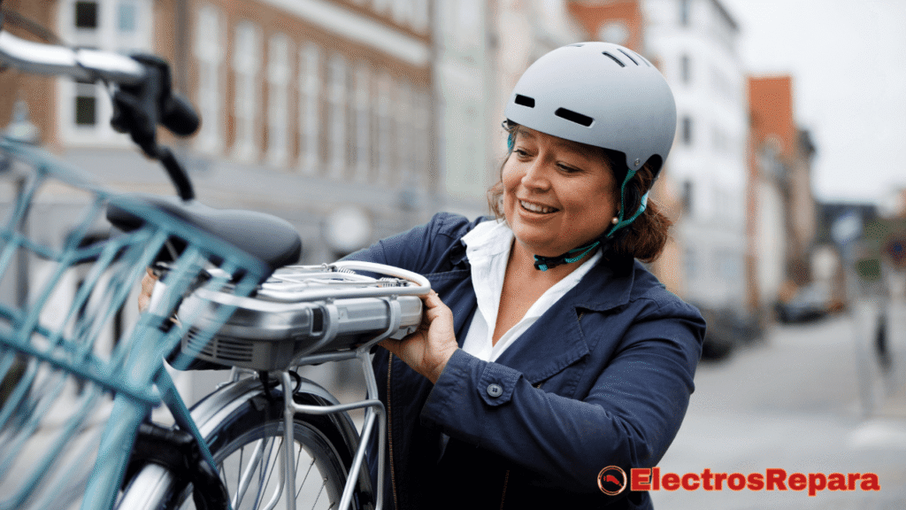 una mujer con una sonrisa, que lleva puesto un casco de ciclista, instalando una batería de bicicleta eléctrica en el portabultos trasero de una bicicleta. La batería es de color plateado y tiene una forma rectangular. En el fondo, se observa una calle de ciudad. 