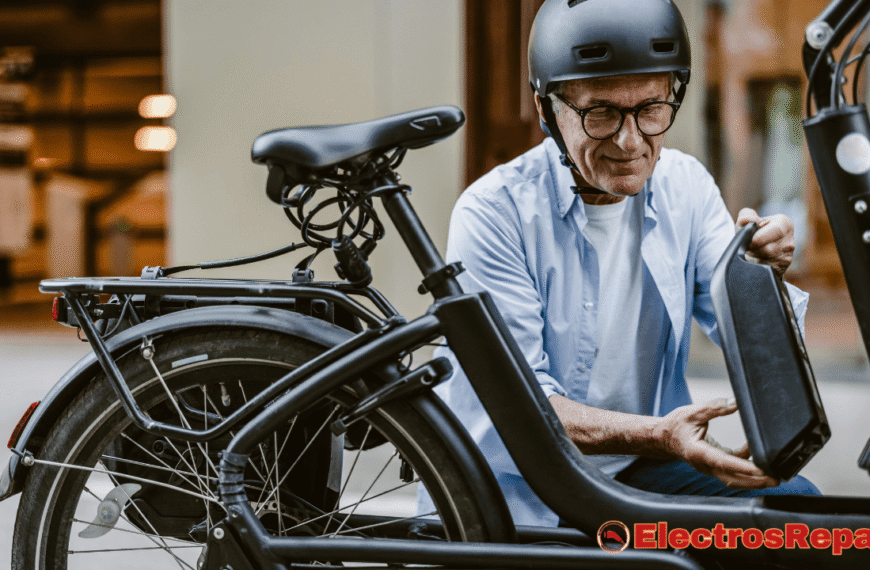 un hombre mayor, que lleva un casco y gafas, instalando o retirando la batería de la bicicleta eléctrica del tubo inferior del cuadro. La batería de la bicicleta eléctrica es un componente de color negro y de forma rectangular. El hombre está inclinado y su mirada está fija en la acción que está realizando. Se puede ver que la bicicleta tiene un diseño moderno con un portabultos trasero. El fondo sugiere un entorno urbano.