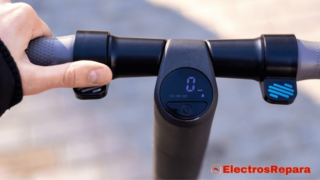 
La imagen muestra un primer plano del manillar de un patinete eléctrico, con una mano sujetando el puño izquierdo, que tiene una textura gris punteada. En el centro del manillar, un display digital muestra un "0" grande y "km/h" debajo, con un indicador de batería con tres barras y un botón de encendido/apagado. A la derecha, se observa una palanca de aceleración o freno con detalles en azul brillante y negro. El manillar es de color negro mate con detalles en gris. En la parte inferior derecha de la imagen, 