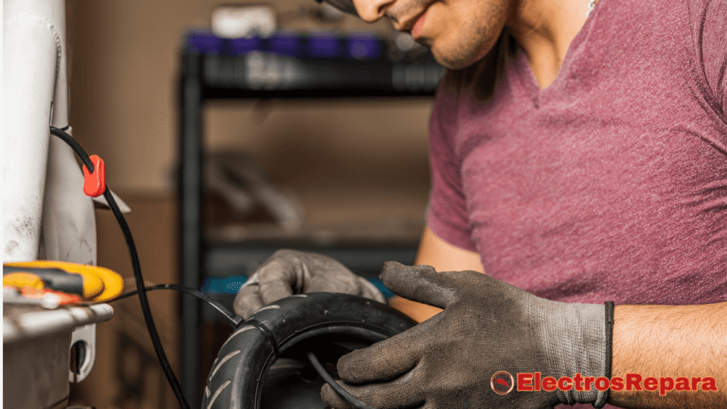 una persona con una camiseta de color vino y guantes de trabajo, manipulando lo que parece ser la rueda de un patinete eléctrico. Se observan cables eléctricos y herramientas en un entorno de taller.