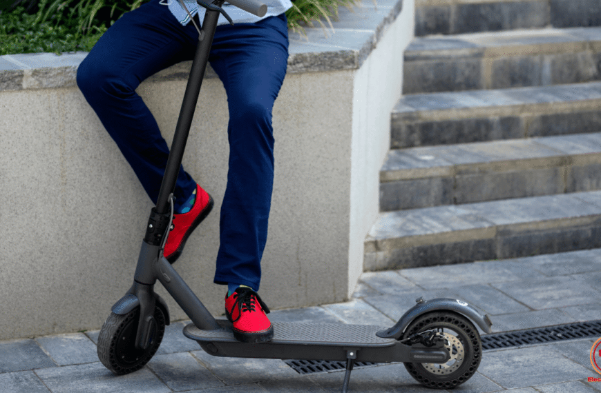 Una persona se encuentra sobre una superficie de piedra con un patinete eléctrico, conectado a la electricidad. La lubricación de las ruedas es crucial para su correcto funcionamiento y mantenimiento.